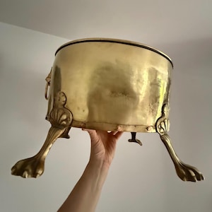 Puede incluir: Un recipiente de metal color latón con un cuerpo redondeado y un borde negro. El recipiente está sostenido por tres patas decorativas con forma de garra. Detalles ornamentados incluyen asas con cabeza de león y adornos decorativos.