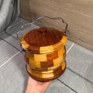 Puede incluir: Un cubo de madera redondo con tapa, que presenta un patrón de tablero de ajedrez con bloques de madera claros y oscuros alternados. El recipiente incluye un asa oscilante de metal plateado en los laterales y un pequeño pomo circular en la tapa.