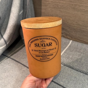May include: A cylindrical, orange-brown ceramic sugar canister with a wooden lid. The canister has a black label with the text "SUGAR" and "The Original Suffolk Canister." Made by Henry Watson Pottery, England, founded 1800.
