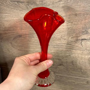 May include: A vibrant red glass vase with a flared top and a slender stem. The vase has a clear, textured base. The hand-blown glass reflects light, creating a glossy appearance. This decorative vase is suitable for floral arrangements.
