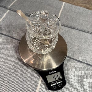 Op de afbeelding: Een heldere, geslepen glazen suikerpot met deksel en een kleine lepel, op een digitale weegschaal. De weegschaal geeft een gewicht van 44,2 aan, waarschijnlijk in gram. De pot staat op een zilverkleurig platform.