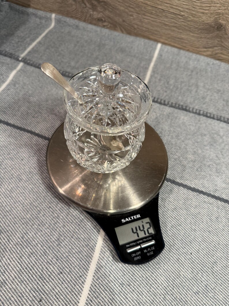Op de afbeelding: Een heldere, geslepen glazen suikerpot met deksel en een kleine lepel, op een digitale weegschaal. De weegschaal geeft een gewicht van 44,2 aan, waarschijnlijk in gram. De pot staat op een zilverkleurig platform.