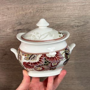以下が含まれることがあります： 白磁の砂糖入れ。蓋付きで、ボルドー、ゴールド、紫色の花柄模様が施されています。ボウルには小さな取っ手と装飾的な縁があります。蓋には小さなつまみが付いています。