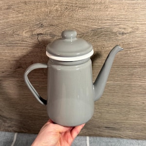 Puede incluir: Una cafetera de esmalte gris con tapa y asa curva. La cafetera tiene un pico largo y un borde blanco alrededor de la tapa. La cafetera está sostenida en la mano de una persona.