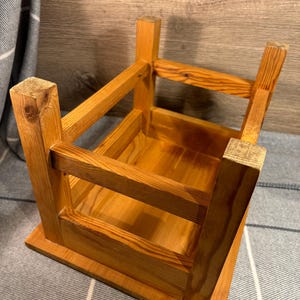 May include: A small, unfinished wooden stool with a square base and a rectangular top. The stool is constructed with visible joints and has a natural wood grain finish. The legs are square and sturdy, and the overall design is simple and rustic.