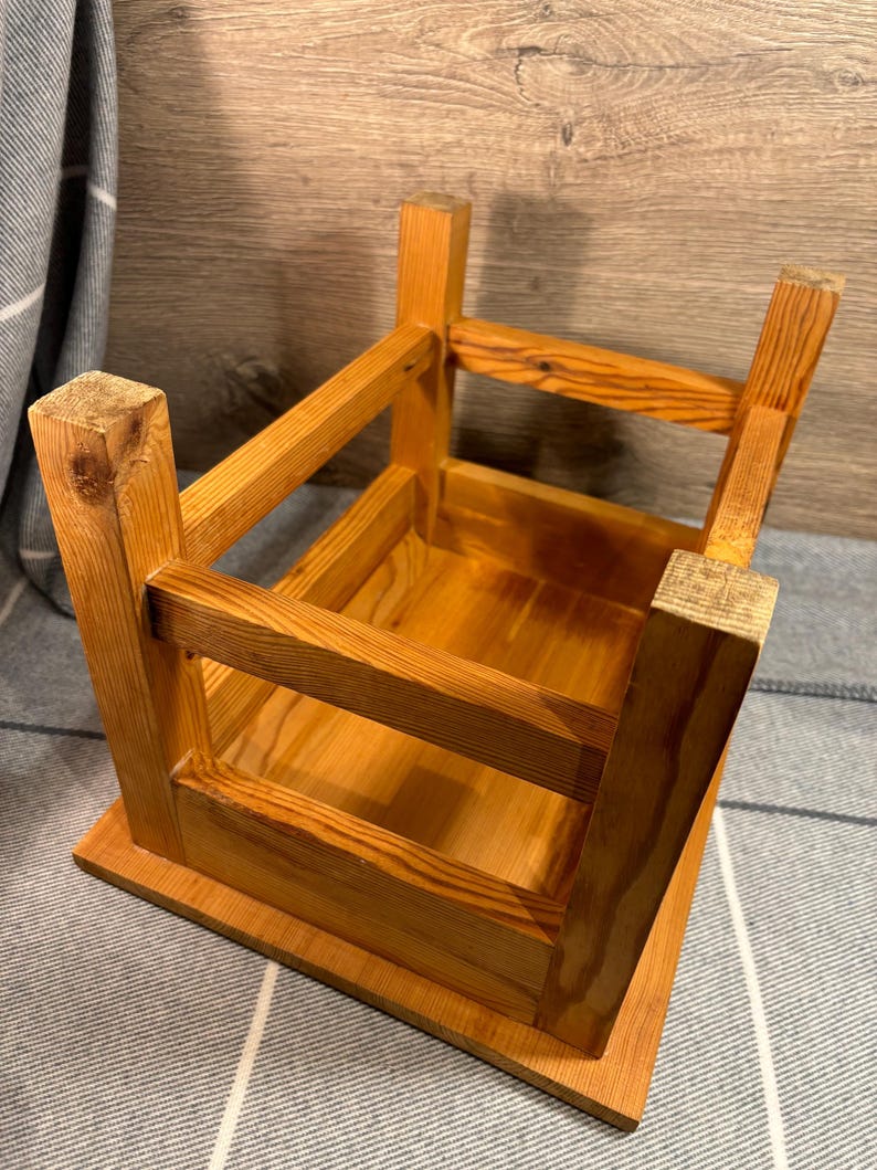 May include: A small, unfinished wooden stool with a square base and a rectangular top. The stool is constructed with visible joints and has a natural wood grain finish. The legs are square and sturdy, and the overall design is simple and rustic.