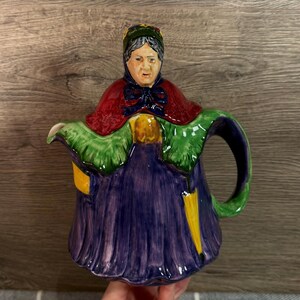 May include: A ceramic teapot shaped like an elderly woman, featuring a red cape, green shawl, and a purple dress. The teapot has a yellow umbrella design and a detailed face, with a green handle and spout.