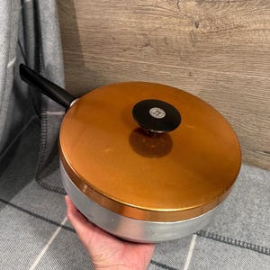May include: A vintage copper-coloured frying pan with a black handle and lid. The pan has a silver-coloured base and a black knob on the lid. The pan is designed for cooking and is likely made of metal.