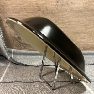 May include: A black, rectangular serving dish with a glossy finish, resting on a clear, angular metal stand. The dish has a light-colored interior and a slightly curved shape. The background is a gray and white patterned fabric.