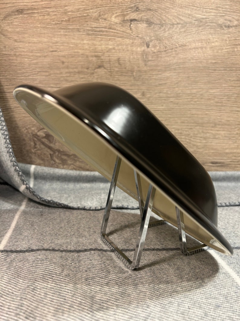 May include: A black, rectangular serving dish with a glossy finish, resting on a clear, angular metal stand. The dish has a light-colored interior and a slightly curved shape. The background is a gray and white patterned fabric.