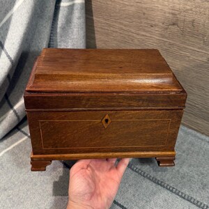 May include: A rectangular wooden box with a hinged lid, featuring a diamond-shaped inlay on the front. The box is a warm brown color with a decorative border and rests on small feet. The box is likely used for storing jewelry or other small items.