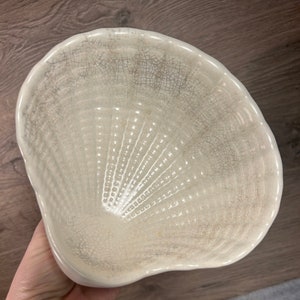 Op de afbeelding: Een witte keramische kom in de vorm van een schelp met een gekrakelde glazuur. De kom heeft een getextureerd, geribbeld interieur.