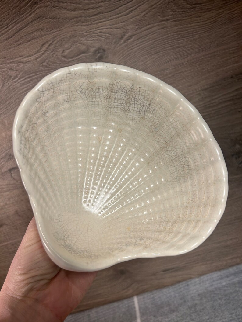 Op de afbeelding: Een witte keramische kom in de vorm van een schelp met een gekrakelde glazuur. De kom heeft een getextureerd, geribbeld interieur.