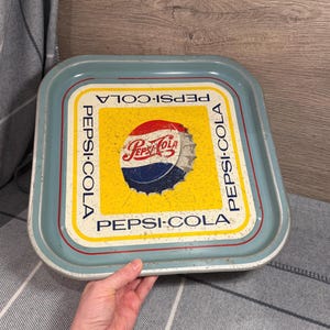 May include: Vintage Pepsi-Cola metal serving tray. The tray has a light blue border with a yellow center featuring the Pepsi logo and bottle cap design. The tray has the words "Pepsi-Cola" printed around the center.