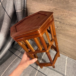 May include: A small, brown bamboo stool with a rectangular top and geometric legs. The stool is made of natural bamboo and has a warm, reddish-brown hue. The top is slightly darker than the legs.