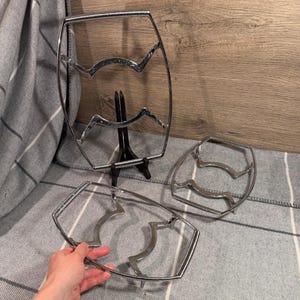 Op de afbeelding: Drie zilverkleurige metalen draadframes met een rechthoekige vorm en gebogen binnensteunen. Eén frame wordt in de hand gehouden, één staat rechtop op een zwarte standaard en één ligt plat. De frames hebben een ontwerp van gedraaid metaaldraad.