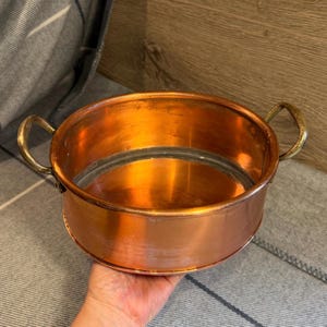 May include: A round copper cooking pot with a polished metallic finish and two curved side handles. The interior of the pot shows a smooth, reflective surface, while the exterior displays a warm, reddish-brown hue consistent with copper metal.