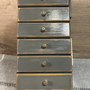 May include: A small wooden dresser with six drawers painted in a dark grey colour. Each drawer has a brass knob and a thin gold trim around the edge.