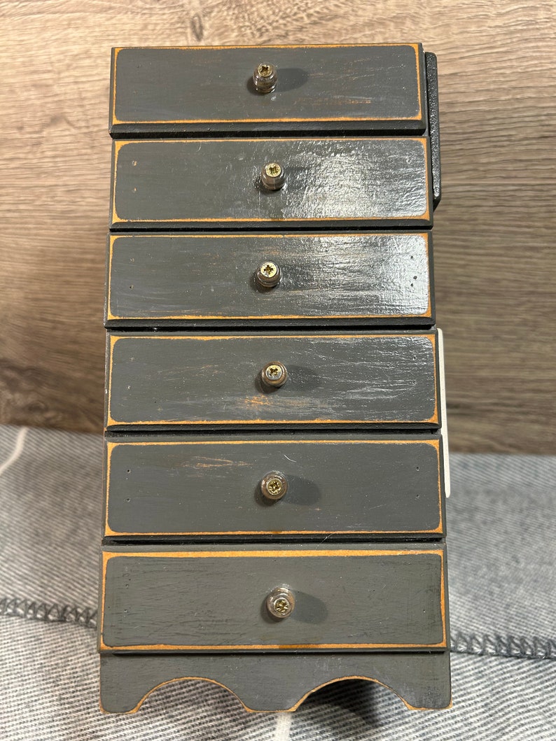 May include: A small wooden dresser with six drawers painted in a dark grey colour. Each drawer has a brass knob and a thin gold trim around the edge.