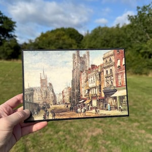 Könnte beinhalten: Eine Vintage-Postkarte mit einer detaillierten Straßenszene mit Gebäuden, Geschäften und Menschen. Das Kunstwerk hat ein klassisches, gealtertes Aussehen mit gedämpften Farben und einem schwarzen Rand. Das Bild wird vor dem Hintergrund von grünem Gras und blauem Himmel gehalten.