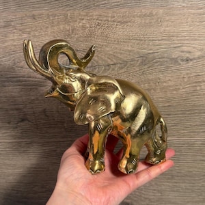 May include: A golden brass elephant figurine with its trunk raised. The elephant has detailed features, including wrinkles and tusks. The figurine is held in a hand, showcasing its size and craftsmanship.