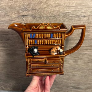 Peut inclure: Pichet en céramique marron en forme de bibliothèque avec une poignée. Le pichet présente une étagère avec des livres miniatures, un téléphone noir et une figurine de chat. Le pichet a un bec verseur.