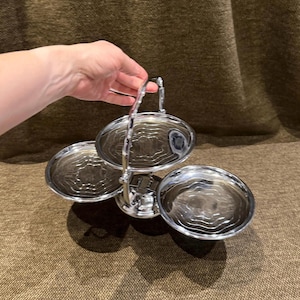 May include: A three-tiered silver-toned metal serving stand featuring three circular trays with a concentric ripple pattern. The stand includes a central vertical handle with a decorative design, allowing for easy transport of the tiered structure.