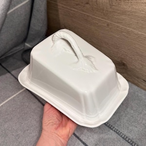 May include: A white ceramic butter dish with a lid and a handle. The handle is curved and has a leaf-like design at the base. The dish is rectangular with rounded corners.
