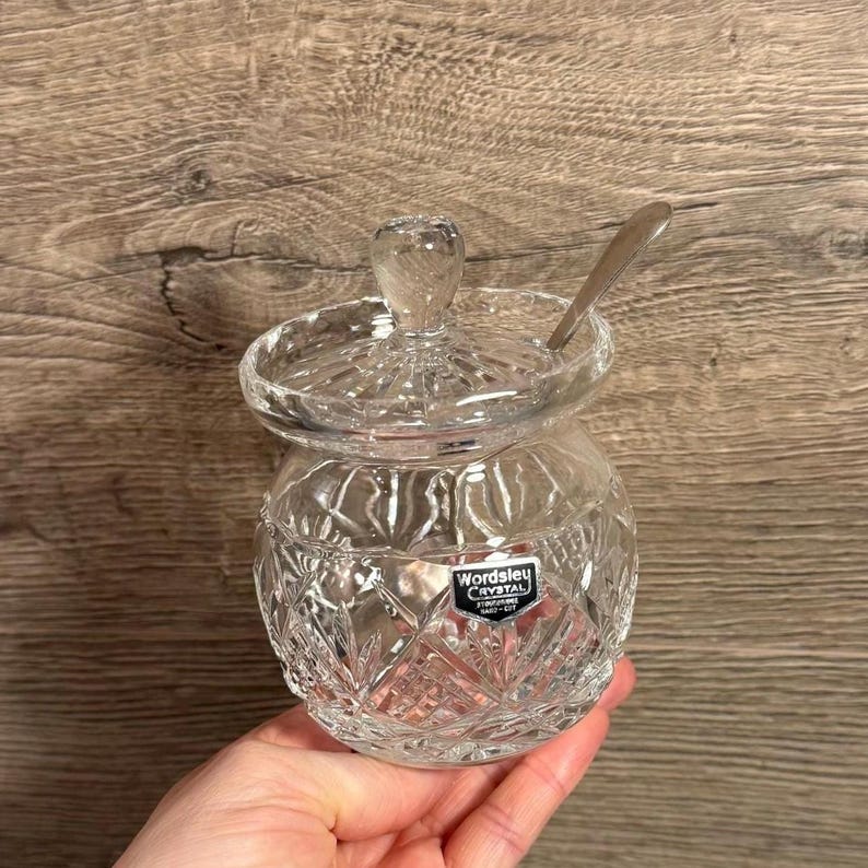 Op de afbeelding: Een heldere kristallen suikerpot met een deksel en een kleine lepel. De kom heeft een diamantgeslepen patroon en een klein handvat op het deksel. Het label Wordsley Crystal is zichtbaar.