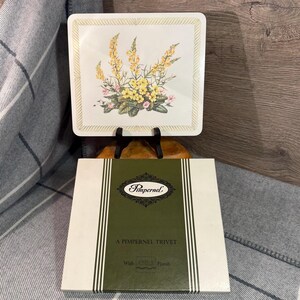 May include: A square trivet with a floral design, featuring yellow and pink flowers, is displayed on a black stand. The trivet is from the brand "Pimpernel" and is in its original packaging.