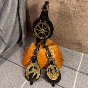 Peut inclure: Ensemble d'attirail de cheval antique, comprenant une pièce de harnais en cuir noir et laiton avec un motif de roue, et deux ornements en cuir et laiton avec des motifs de cheval et de cavalier. Les articles sont exposés sur une base en bois.