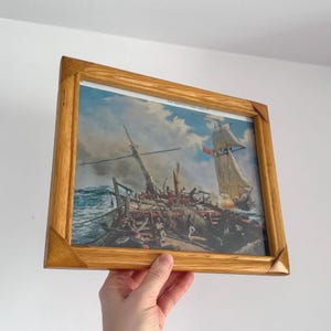 May include: A framed print of a naval battle scene, featuring two sailing ships on a turbulent sea. The print is encased in a light brown wooden frame. The artwork shows a ship in distress and another ship with sails billowing.