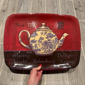 Può includere: Vassoio rettangolare con sfondo rosso e illustrazione decorativa di teiera. La teiera è gialla e blu con un design di ispirazione asiatica. Le parole "Black TEA" sono in alto.