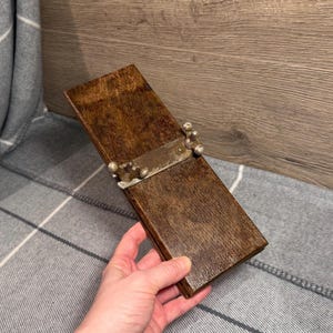 May include: A vintage wooden box with a hinged lid and metal hardware. The box is rectangular and made of dark brown wood. The metal hardware is silver-colored and includes screws and decorative elements. The box is held by a person's hand.