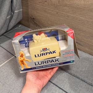 Könnte beinhalten: Eine Lurpak Butterdose in der Originalverpackung aus Karton. Die Schachtel zeigt eine Grafik der Figur Douglas und Text, einschließlich Limited Edition Douglas Butter Dish und 2 x 250g. Die Butterdose ist cremefarben mit dem Lurpak-Logo.