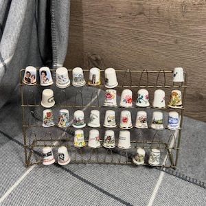 Puede incluir: Una colección de dedales vintage expuestos en un estante de metal dorado. Los dedales presentan varios diseños, incluyendo patrones florales, retratos y vistas panorámicas. El estante está colocado sobre una superficie con estampado gris y blanco.