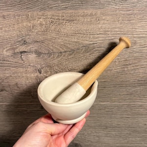 Peut inclure: Un petit ensemble mortier et pilon de couleur blanc cassé. Le mortier a la forme d'un bol arrondi, et le pilon est en bois clair. Le pilon repose à l'intérieur du mortier. L'ensemble est conçu pour broyer les herbes et les épices.
