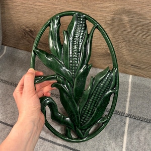 May include: Green ceramic corn on the cob hot pad. The hot pad is shaped like an oval and has a raised design of two ears of corn with leaves.