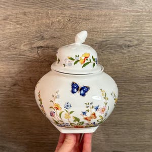 Op de afbeelding: Een witte keramische pot met deksel, versierd met kleurrijke bloemen- en vlindermotieven. De pot heeft een afgeronde body en een deksel met een kleine, witte knop. De bloempatronen omvatten gele, blauwe en oranje bloemen.