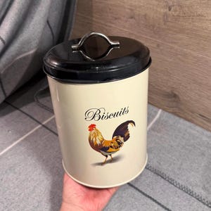 May include: A cream-colored cylindrical biscuit canister with a black lid and a silver metal handle. The front features a colorful rooster illustration and the word Biscuits in a black script font. The container has a smooth finish.