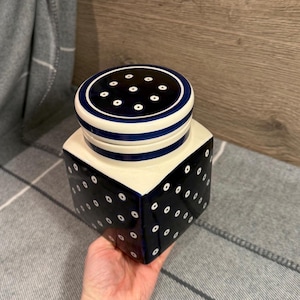Op de afbeelding: Een keramische pot met deksel, in marineblauw en crème. De kubusvormige pot heeft witte stippenpatronen op de marineblauwe zijkanten. Het deksel is marineblauw met witte stippen en blauwe strepen. De pot wordt door een hand vastgehouden.