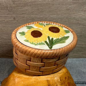 Può includere: Un portagioie in ceramica a forma di cestino, con un coperchio decorato con girasoli. Il coperchio mostra due girasoli gialli con centri marroni e foglie verdi. Il cestino è marrone chiaro con una trama intrecciata.