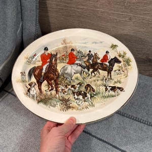 Puede incluir: Plato ovalado para servir con una escena de caza. El plato crema presenta un borde dorado y una ilustración detallada de jinetes a caballo, perros y un paisaje campestre. Los jinetes visten abrigos rojos.