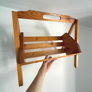 May include: A light brown wooden magazine rack with a handle at the top. The rack has a slatted design and is held up by two legs. The wood grain is visible. The rack is being held up by a person's hand.