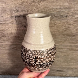 Puede incluir: Un jarrón de cerámica beige con un acabado moteado. La parte inferior presenta un diseño marrón detallado. El jarrón tiene una base ancha y una abertura ligeramente ensanchada. El jarrón es sostenido por una mano.