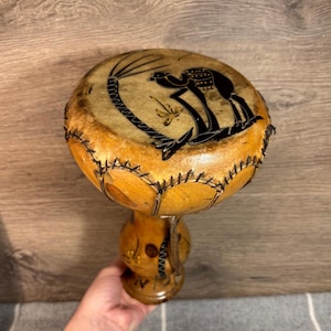 May include: A small, wooden drum with a brown leather head. The head is decorated with a black silhouette of a camel and a palm tree. The drum has a wooden base with a carved design.