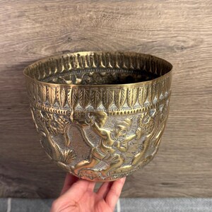 Op de afbeelding: Een messing kom met ingewikkelde reliëfsnijwerk. De kom heeft gedetailleerde figuren en patronen rond de buitenkant. De rand heeft een decoratieve rand. De kom wordt door een hand vastgehouden.
