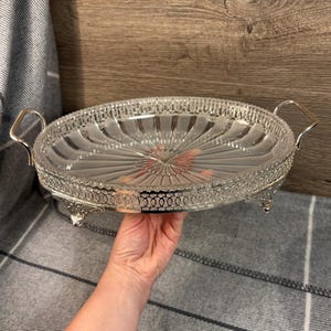 Puede incluir: Una bandeja de servir plateada con un inserto de vidrio transparente. La bandeja tiene un borde decorativo y dos asas. El inserto de vidrio tiene un diseño acanalado.