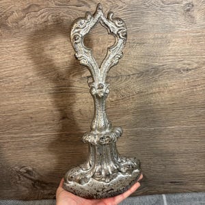 May include: A silver metal doorstop with an ornate design. The doorstop is shaped like a key with a large, decorative head and a smaller, round base.