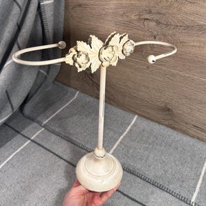 May include: Off-white metal towel holder with a floral design. The holder has a round base, a central stem, and two curved arms. The floral design is centered on the top of the stem. The holder has a distressed, aged finish.